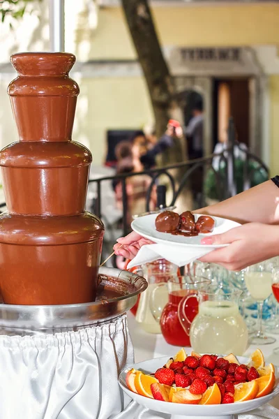 Chocolate fondue with fruits assortment. Female hand dipping strawberry on a skewer into the warm chocolate fondue fountain at the party. Catering food. — Stock Photo, Image