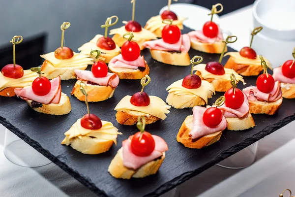 Small crostini with grilled baguette, cherry tomatoes, ham slices, cheese and fresh grapes on black background. Sandwiches or assorted canapes on a dark board, top view. — Stock Photo, Image