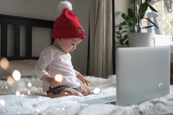 Bebé en sombrero de Navidad charlando a través de lap top en casa durante la cuarentena. Distanciamiento social durante el bloqueo. Año nuevo y la celebración de Navidad en casa — Foto de Stock