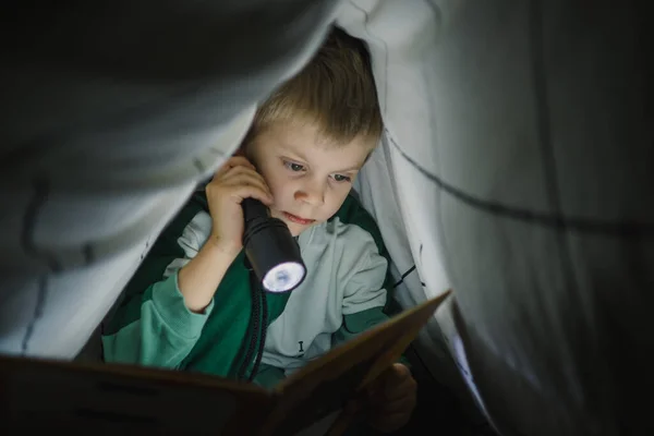 Küçük çocuk geceleri battaniyeyle kaplı bir fenerle kitap okuyor. — Stok fotoğraf