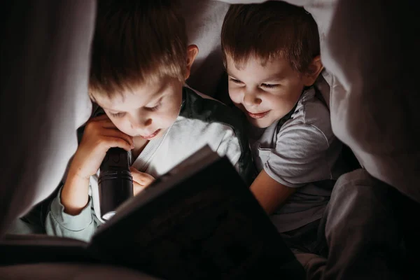 Два младших брата ночью читают книгу, прячась под одеялом. — стоковое фото