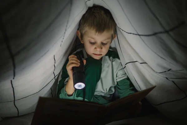 Küçük çocuk geceleri battaniyeyle kaplı bir fenerle kitap okuyor. — Stok fotoğraf