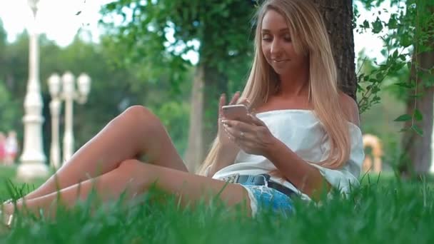 Une jeune fille tenant un smartphone est assise sur l'herbe verte du parc de la ville. Fille porte un t-shirt blanc . Clip Vidéo