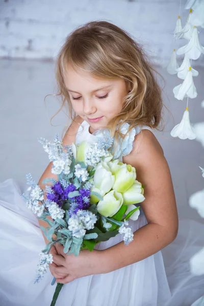 Piękne blond dziewczynka kaukaski siedzi z bukietem wiosennych kwiatów w białej sukni. — Zdjęcie stockowe