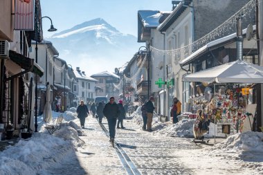 Bansko, Bulgaristan - 17 Ocak 2025: İnsanlar Avrupa 'daki bir dağ tatil beldesinde, karla kaplı dükkanlar ve görkemli bir dağ zirvesi ile canlı, güneşli bir yaya caddesinde yürüyor.