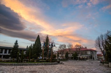 Canlı bir günbatımı, geleneksel binalar, yüksek yeşil ağaçlar ve dramatik bulutların altında asfalt bir toplanma alanı içeren tarihi kent meydanında sıcak renkler bırakıyor..