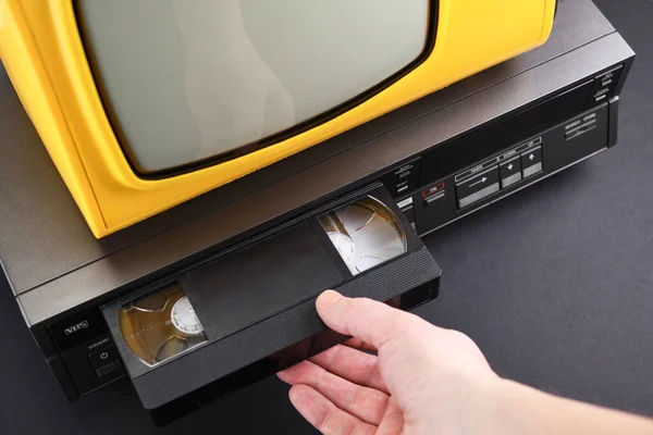 VHS videocassette is put into the video recorder to watch the video. Old yellow vintage TV with VCR and videotape on black background from 1980s, 1990s, 2000s.