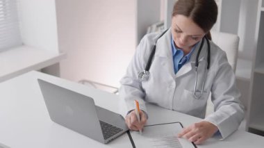 Beyaz önlüğüyle, kendini adamış bir kadın doktor hasta notlarını dizüstü bilgisayarında inceleyerek, sağlık, telgraf ve genel sağlıktaki modern gelişmelerin önemini vurguluyor.