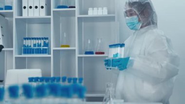 Çağdaş bir laboratuvar ortamında, gerekli koruyucu malzemelerle süslenmiş çalışkan bir laboratuvar bilimcisi, önemli araştırma girişimlerini temsil eden çeşitli test tüplerini titizlikle inceliyor.