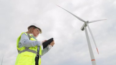 Güvenlik teçhizatı alanında yetenekli bir kadın mühendis, sürdürülebilir bir geleceğin geliştirilmesinde hayati rol oynayan yenilenebilir enerjileri ön plana çıkaran bir rüzgar türbini alanındaki faaliyetleri izlemek için bir tablet kullanıyor.