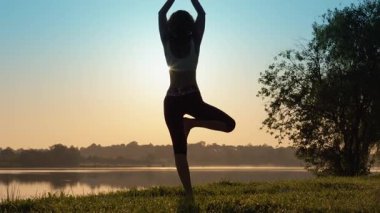 Gün doğumunda yoga yapan huzurlu bir kadın silueti, mükemmel bir şekilde somutlaştırıcı sükunet ve denge, güzel manzaralı göl kenarı zeminine karşı, refah ve uyumu teşvik eder.