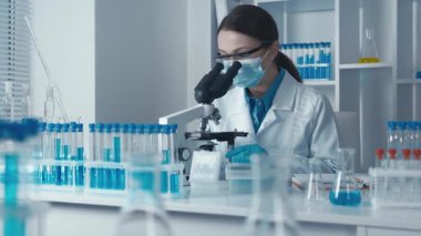 Odaklanmış bir kadın bilim adamı mikroskop kullanarak araştırmalarını yürütüyor. Etrafı renkli test tüpleri ve modern laboratuvar ortamlarıyla çevrili. Bilimsel araştırmalarda kendini adamışlık ve hassasiyet sergiliyor..