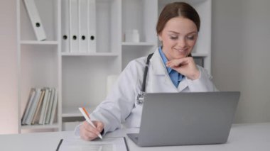 Laboratuvar önlüğü giymiş kendini adamış bir sağlık çalışanı laptopla düşünceli bir şekilde etkileşime girer, notlar alır ve bilgileri inceler, dijital sağlık hizmetlerine modern değişimi somutlaştırır..