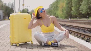 Canlı, sarı elbiseli şık bir genç kadın, bir tren platformunda aynı bavulla oturur ve neşe ve keşiflerle dolu heyecanlı bir yaz yolculuğunu heyecanla bekler.