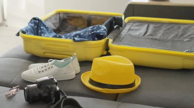 Parlak sarı bir bavul, içi seyahat gereksinimleriyle dolu iyi korunmuş bir iç mekan gösteriyor. Yakınlarda, taze beyaz ayakkabılar, parlak sarı bir şapka ve önümüzde heyecan verici maceraları bekleyen bir kamera.