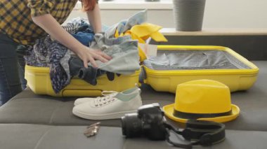 Kişi titizlikle parlak sarı bir bavulu giysi ve seyahat eşyalarıyla dolduruyor. Etrafı bitkilerle çevrili ve tatil hazırlığı için ideal bir ortam.