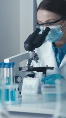 Odaklanmış bir kadın bilim adamı mikroskop kullanarak araştırmalarını yürütüyor. Etrafı renkli test tüpleri ve modern laboratuvar ortamlarıyla çevrili. Bilimsel araştırmalarda kendini adamışlık ve hassasiyet sergiliyor..