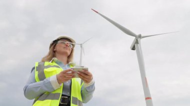 Güvenlik teçhizatında yetenekli bir kadın mühendis, yüksek rüzgar türbinleri yakınında bir İHA çalıştırıyor, yenilenebilir enerjide teknolojinin hayati rolünü gösteriyor ve sürdürülebilirlik girişimlerini destekliyor.