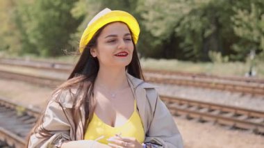 Parlak, güneşli, sarı bir kıyafet giymiş genç bir kadın yaklaşan yaz macerası için heyecanla dolu bavulunu şık bir şekilde elinde, tren rayları boyunca güvenle geziniyor.