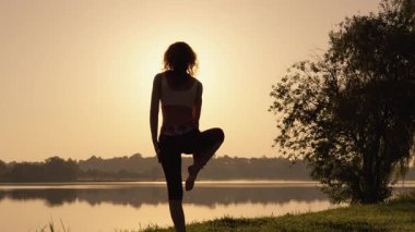 Sabah ışığında, bir kadın huzur ve uyumu somutlaştıran sakin bir gölün yanında yoga yapıyor, doğayla derin bir bağ kuruyor, seyircilere hareketleriyle ve sakin tavırlarıyla ilham veriyor.