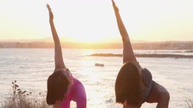 Kadınlar deniz kenarında sakin bir yoga seansının keyfini çıkarırlar. Güneş batarken zarif bir şekilde uzanırlar. Huzurlu manzara ve senkronize hareket denge ve farkındalığı çağrıştırıyor