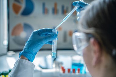 Bir laboratuvar çalışanı pipet ve test tüpü kullanarak hassasiyet ve güvenliğe odaklanan bir kimyasal deney gerçekleştirir. Bilimsel veri ve araştırma grafikleri araştırmacının arkasında görüntülenir