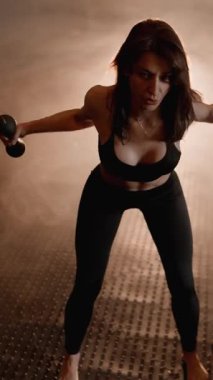 Atletik kadın dinamik bir egzersize girişiyor, dambılları tutuyor ve desenli spor salonunda güçlü bir duruş sergiliyor. Sıcak arka plan onun kendini adamışlığını ve enerjisini vurguluyor.
