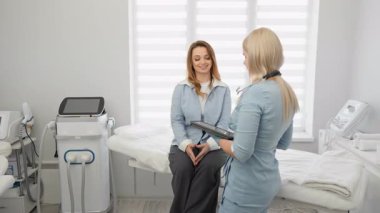 Kadın bir sağlık uzmanı, iyi donanımlı bir klinik odasında bir kadın hastayla tıbbi bilgileri tartışıyor. Sahne güven, modern teknoloji ve kişisel tıbbi hizmeti vurguluyor