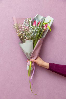 Mor kazaklı bir el, menekşe bir duvarın arka planına karşı bir paket içinde bir buket tutar.