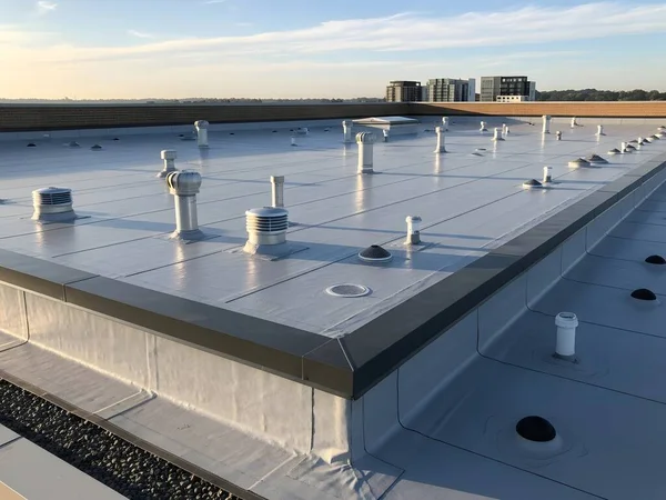 Çeşitli havalandırma sistemlerine sahip modern bir ticari binanın çatısı ve arka planda kentsel mimarinin ve endüstriyel tasarımın sergilendiği bir şehir manzarası..