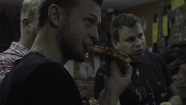Мужчины смотрят, слушают и едят пиццу. Группа людей, ужинающих фаст-фудом в комнате — стоковое видео
