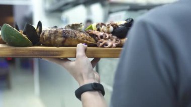 Garson, ahşap tepsi ve ızgara deniz yemekleri taşıyor.