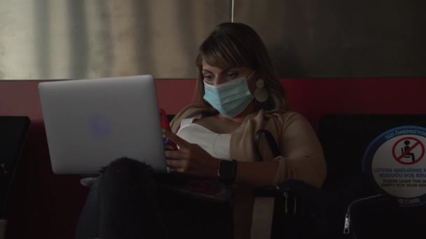 Жінка в масці з ноутбуком і телефоном чекає на політ в залі — стокове відео