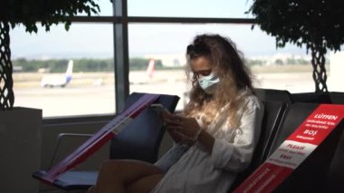 Maskeli kadın havaalanı salonunda cep telefonu kullanıyor.