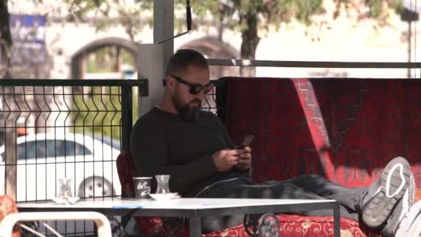 Facet w brodzie i okularach siedzi na czerwonej kanapie w kawiarni. — Wideo stockowe
