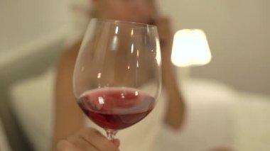 Beyaz havluyla bulanık genç bir kadın yatakta kırmızı şarap tadıyor.