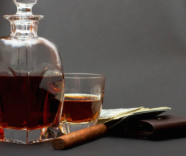 Бутылка с виски и стакан с виски, сигарой и пачкой денег на коричневом бумажнике на темном фоне — стоковое фото