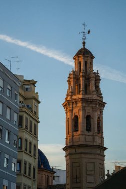Sıcak akşam ışığı altında şehir binalarının üzerinde yükselen tarihi bir kilise kulesi. Dikey mimari şehir manzarası.