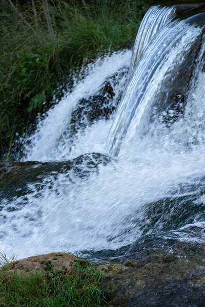 A small cascading waterfall flowing over rocks surrounded by grass and natural vegetation. Vertical outdoor landscape scene.