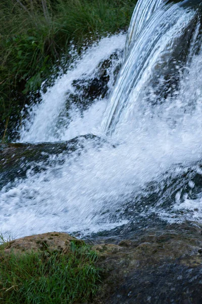 A small cascading waterfall flowing over textured rocks surrounded by green vegetation. Vertical outdoor landscape scene with natural motion.