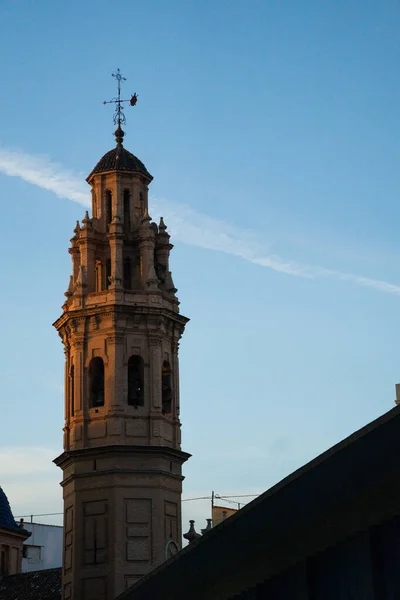 A historic church tower illuminated by warm sunset light, standing against a clear blue sky with soft cloud streaks. Vertical architectural cityscape.