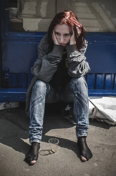 Сумна дівчина в старому рваному брудному одязі і взутті сидить на тротуарі і є порожньою пластиковою чашкою перед нею. Бездомних. Бідність — стокове фото