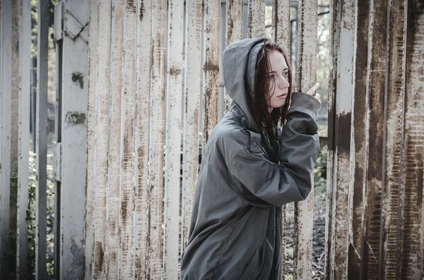 Menina triste na velha roupa suja de pé na cerca de ferro e, infelizmente, olha através dele. Devshuka sem-abrigo. Um adolescente sem-teto . — Fotografia de Stock