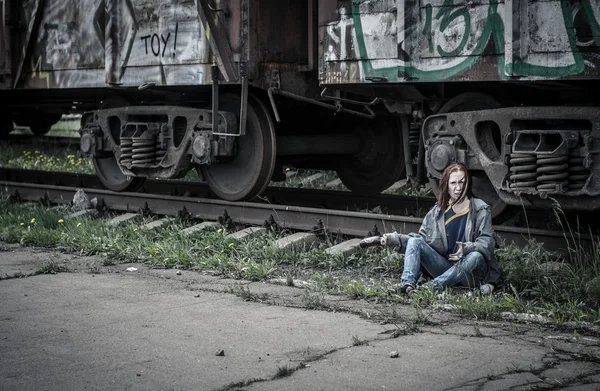 Homeless woman in old, torn, dirty clothes, sitting on the ground next to the train on the railway svagonom and aggressive swings on someone an empty glass bottle — Stock Photo, Image