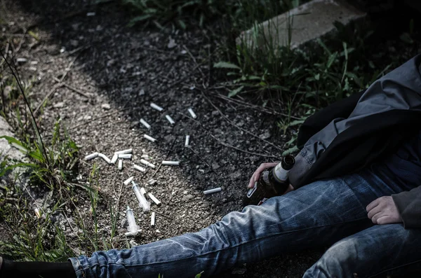 Vieil homme en vêtements déchirés et sales assis sur le sol près de la voie ferrée et tenant une bouteille d'alcool à la main, autour de la sigaretyi dispersés seringues utilisées, aiguilles Photo De Stock