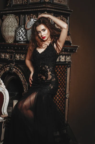 Beautiful slender brunette woman in a long black lace dress stands near the ancient fireplace — Stock Photo, Image