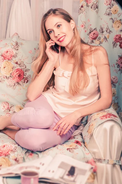 Όμορφη χαρούμενοι νεαρή ξανθιά κοπέλα που κάθεται στον καναπέ μιλάτε στο τηλέφωνο, και χαμογελαστός — Φωτογραφία Αρχείου