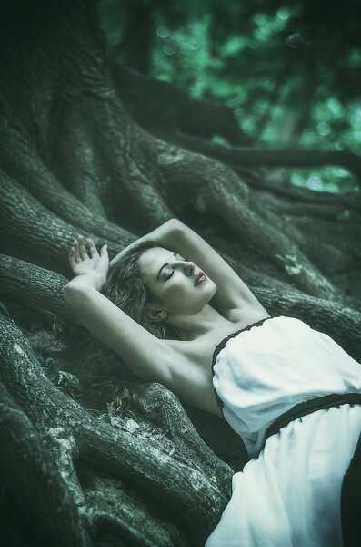 Beautiful young woman in a long white dress with long wavy hair, lying on the large roots of a tree with his eyes closed Fairy tale. Dream