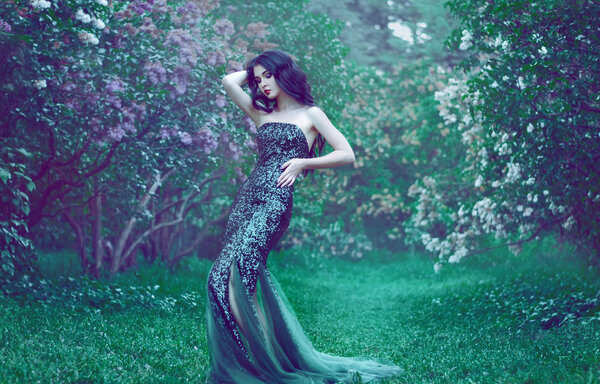Pretty slim girl with dark hair in a long emerald green dress with bare shoulders is in the garden with blooming lilac bushes