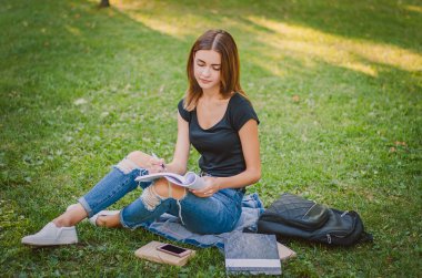 genel bir parkta çim üzerinde oturan ve yanında bir kitap okuma odaklı kız öğrenci bir defter, kalem ve sırt çantası koymak